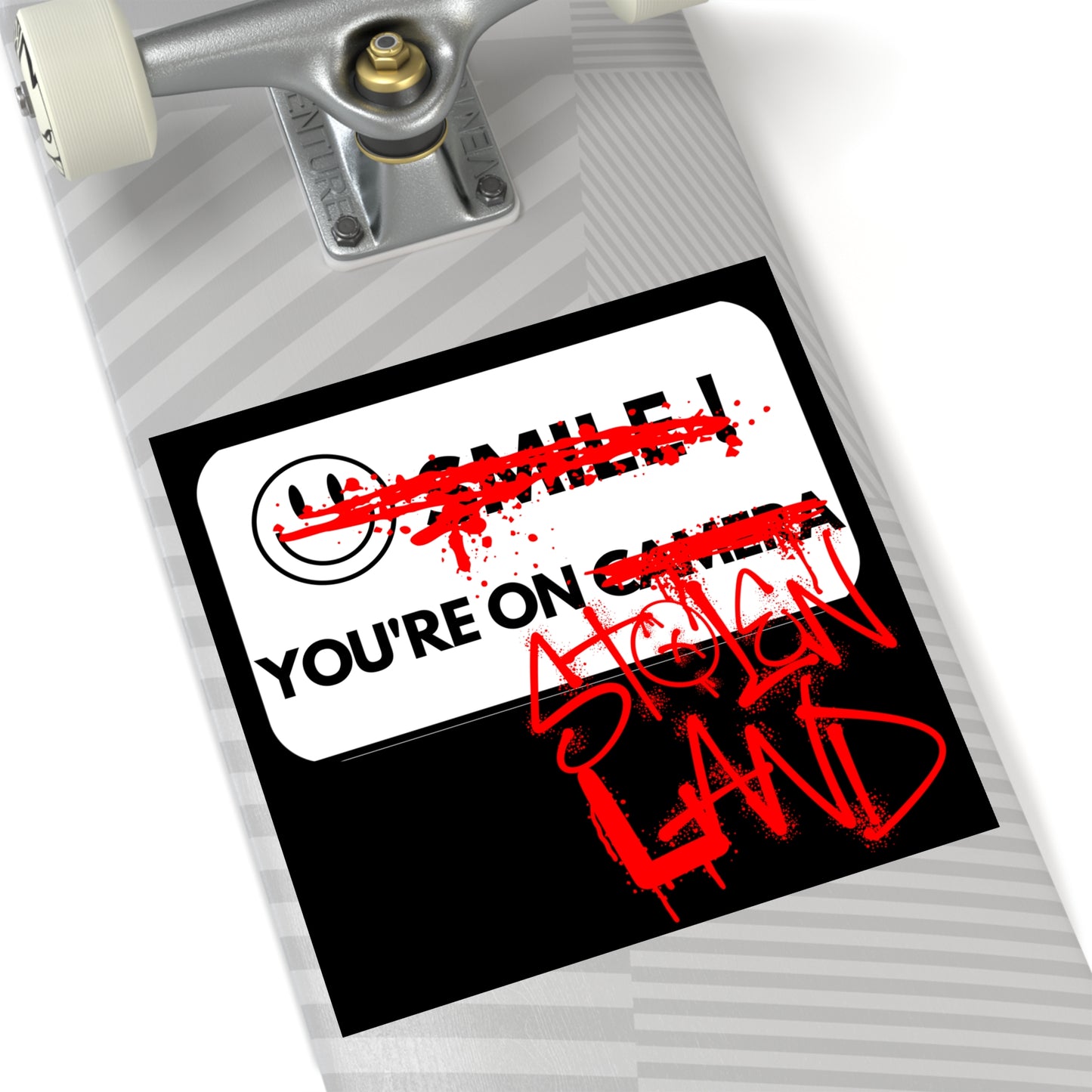 A BLACK STICKER FEATURING A WHITE "SMILE YOU'RE ON CAMERA" SIGN WHERE THE SMILEY FACE AND WORDS "SMILE" AND "CAMERA" HAVE BEEN GRAFFITIED OUT WITH RED SPRAY PAINT AND THE WORDS "STOLEN LAND" HAVE BEEN ADDED AFTER "YOU'RE ON" IN RED SPRAY PAINT