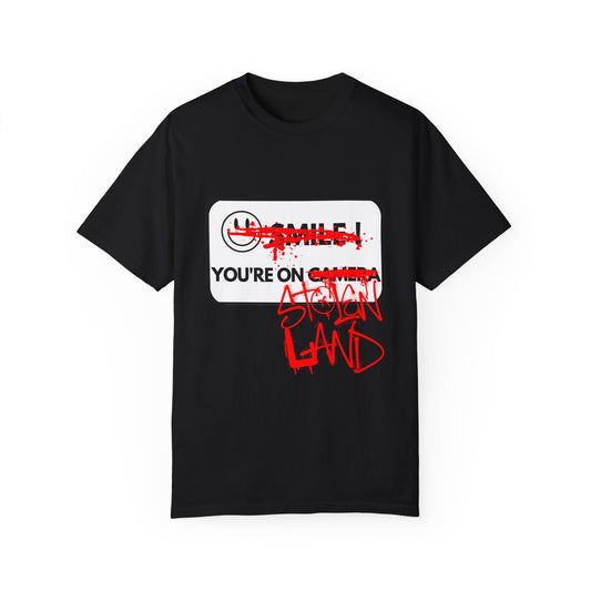 A BLACK T-SHIRT FEATURING A WHITE "SMILE YOU'RE ON CAMERA" SIGN WHERE THE SMILEY FACE AND WORDS "SMILE" AND "CAMERA" HAVE BEEN GRAFFITIED OUT WITH RED SPRAY PAINT AND THE WORDS "STOLEN LAND" HAVE BEEN ADDED AFTER "YOU'RE ON" IN RED SPRAY PAINT